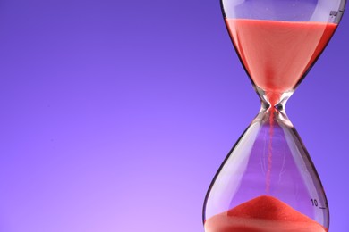 Hourglass with red flowing sand on purple background, closeup. Space for text
