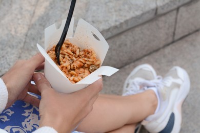 Photo of Woman holding paper box of takeaway noodles with fork outdoors, closeup. Street food