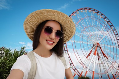 Smiling young woman in sunglasses and straw hat taking selfie near observation wheel