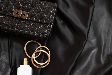 Photo of Leather bag, bottle of perfume and golden bracelets on black fabric, flat lay. Space for text