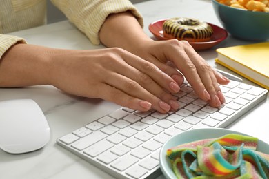 Bad eating habits. Woman working on computer at white marble table with different snacks, closeup