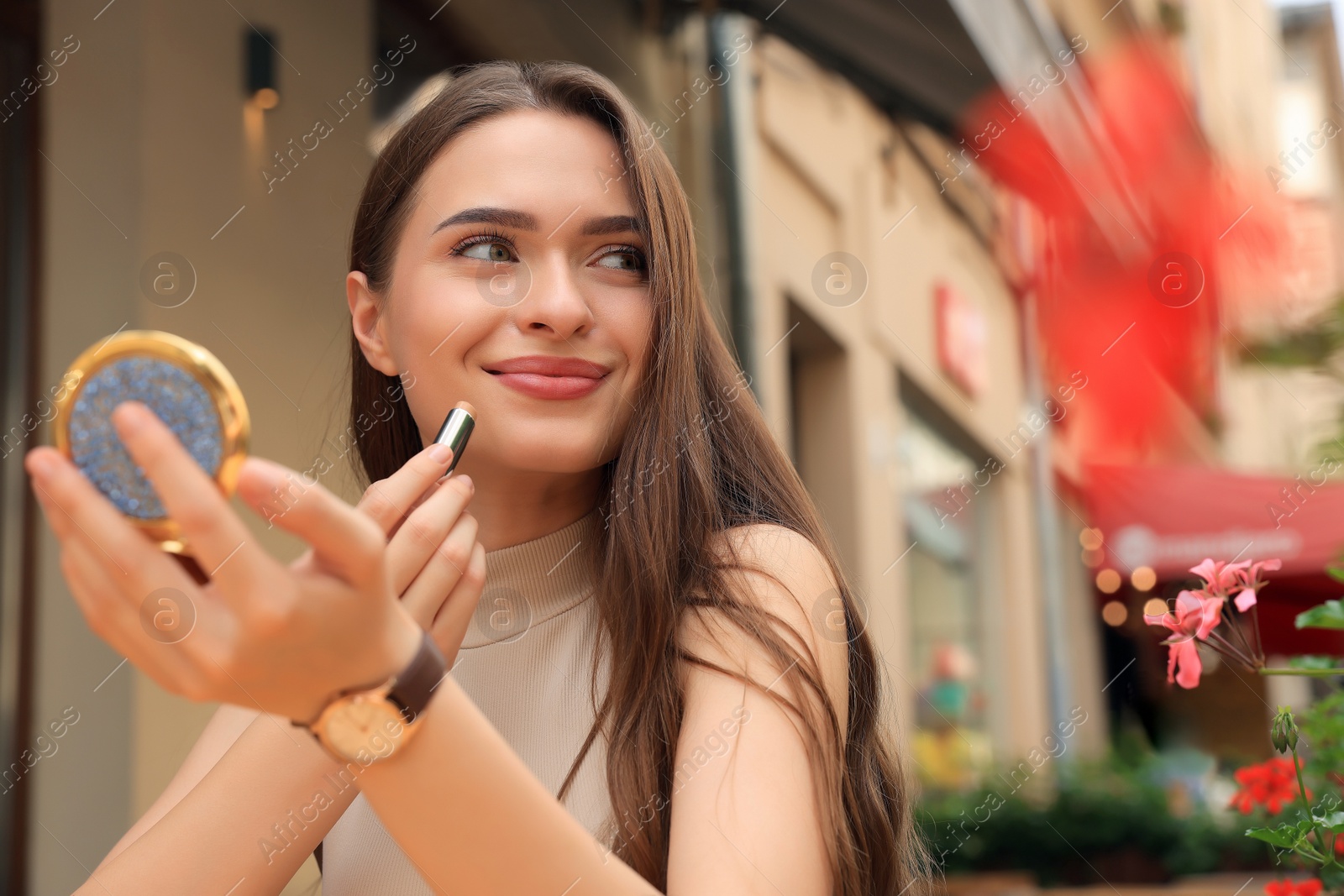 Photo of Beautiful young woman with cosmetic pocket mirror applying lipstick outdoors, space for text