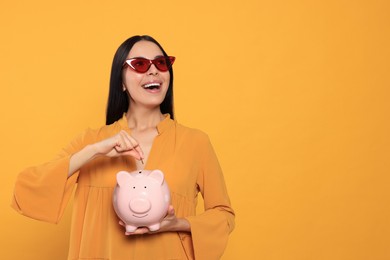 Photo of Emotional young woman putting coin into piggy bank on orange background. Space for text