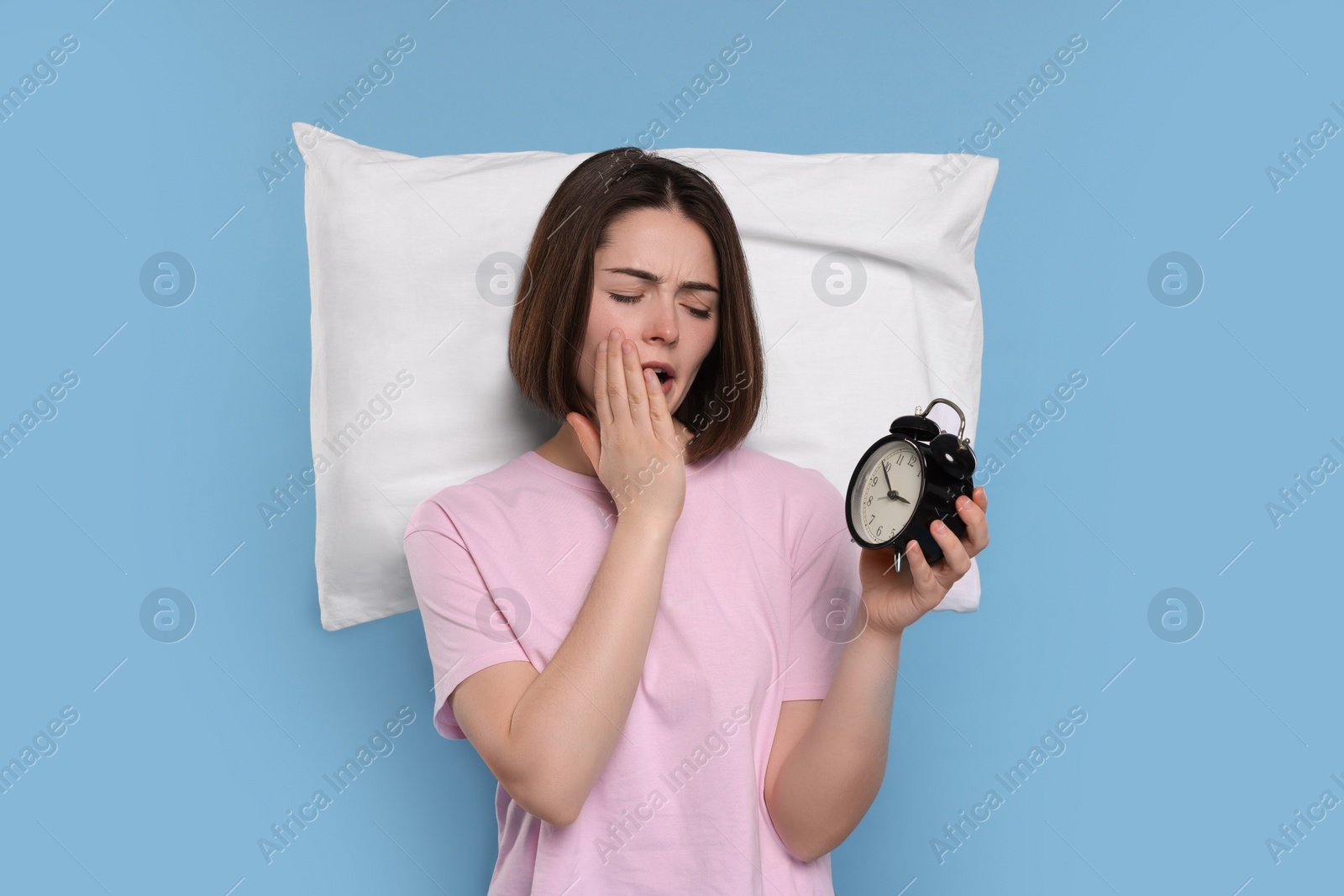 Photo of Sleepy young woman with pillow and alarm clock yawning on light blue background. Insomnia problem