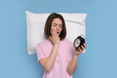 Photo of Sleepy young woman with pillow and alarm clock yawning on light blue background. Insomnia problem