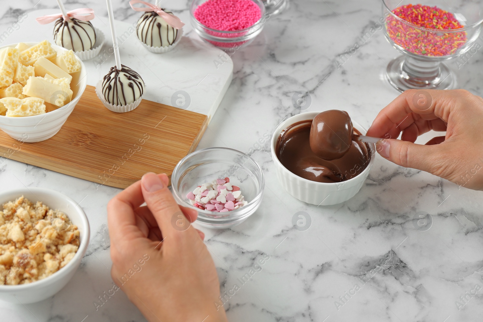 Photo of Young woman putting cake pop into chocolate frosting at white marble table, closeup