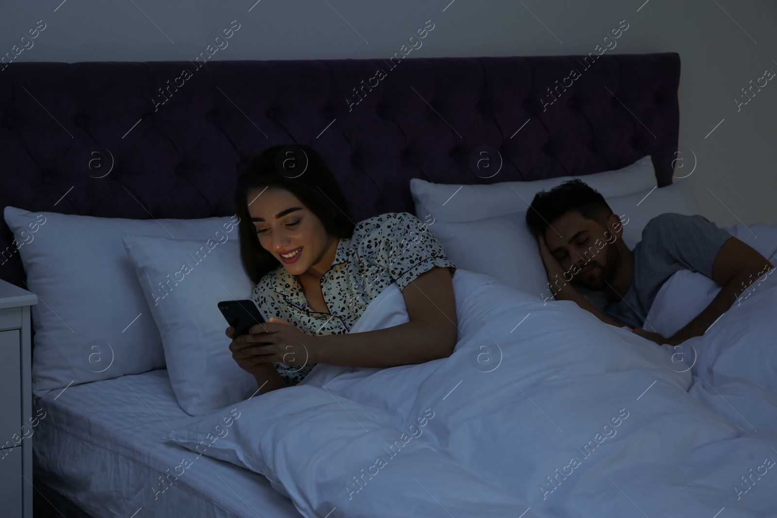 Photo of Young woman using smartphone while her boyfriend sleeping in bed at night