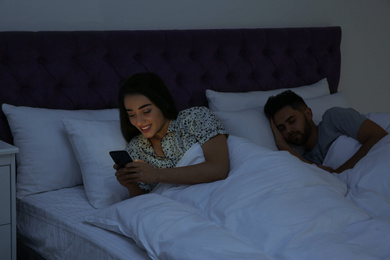 Photo of Young woman using smartphone while her boyfriend sleeping in bed at night
