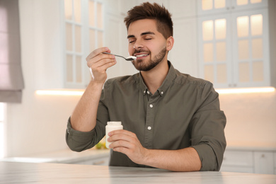 Happy young man eating tasty yogurt at table in kitchen