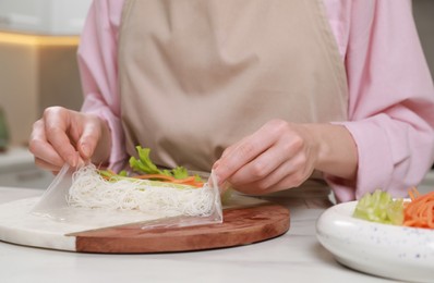 Photo of Making delicious spring rolls. Woman wrapping ingredients into rice paper at white table, closeup