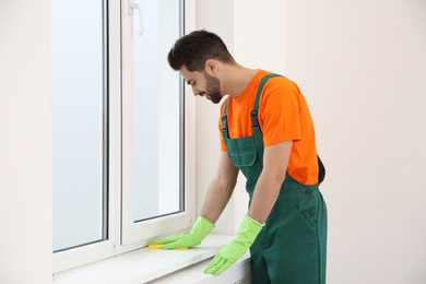 Professional young janitor cleaning windowsill in room