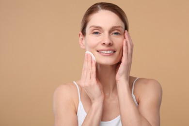 Beautiful woman removing makeup with cotton pad on beige background, space for text