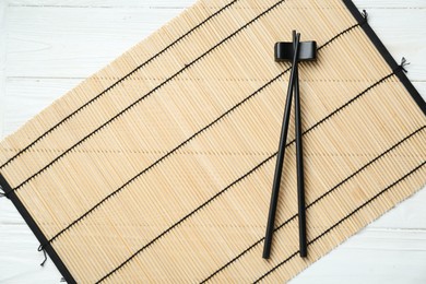 Photo of Bamboo mat with pair of black chopsticks and rest on white wooden table, top view