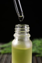 Photo of Dripping dill essential oil from pipette into bottle against black background, closeup