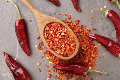 Photo of Chili pepper flakes, pods and spoon on grey table, flat lay