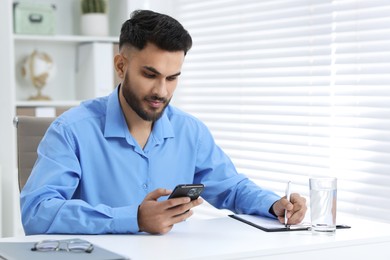 Photo of Handsome young man using smartphone while working at white table in office