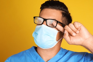 Photo of Doctor wiping foggy glasses caused by wearing medical mask on yellow background, closeup