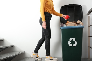 Photo of Young woman throwing coffee cup in trash bin indoors, closeup. Waste recycling