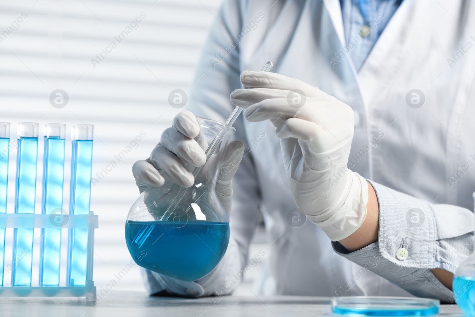 Photo of Scientist taking sample of light blue liquid at white table in laboratory, closeup