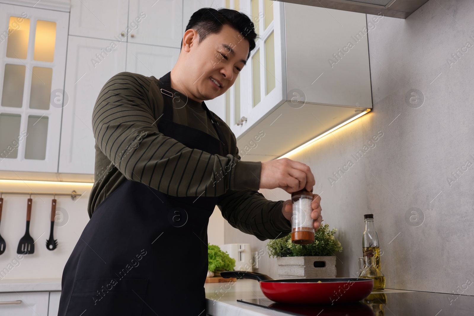 Photo of Cooking process. Man adding salt into frying pan in kitchen, low angle view