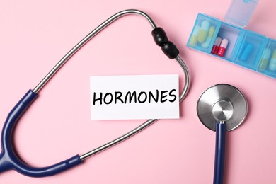 Sheet of paper with word Hormones, stethoscope and pills on pink background, flat lay