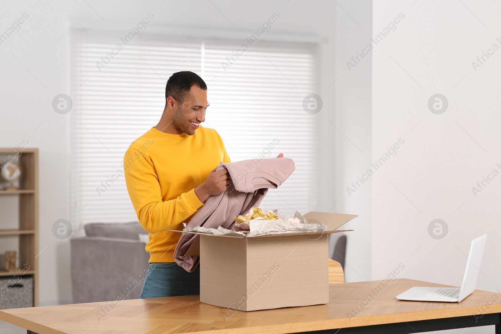 Photo of Happy young man opening parcel at table indoors. Internet shopping