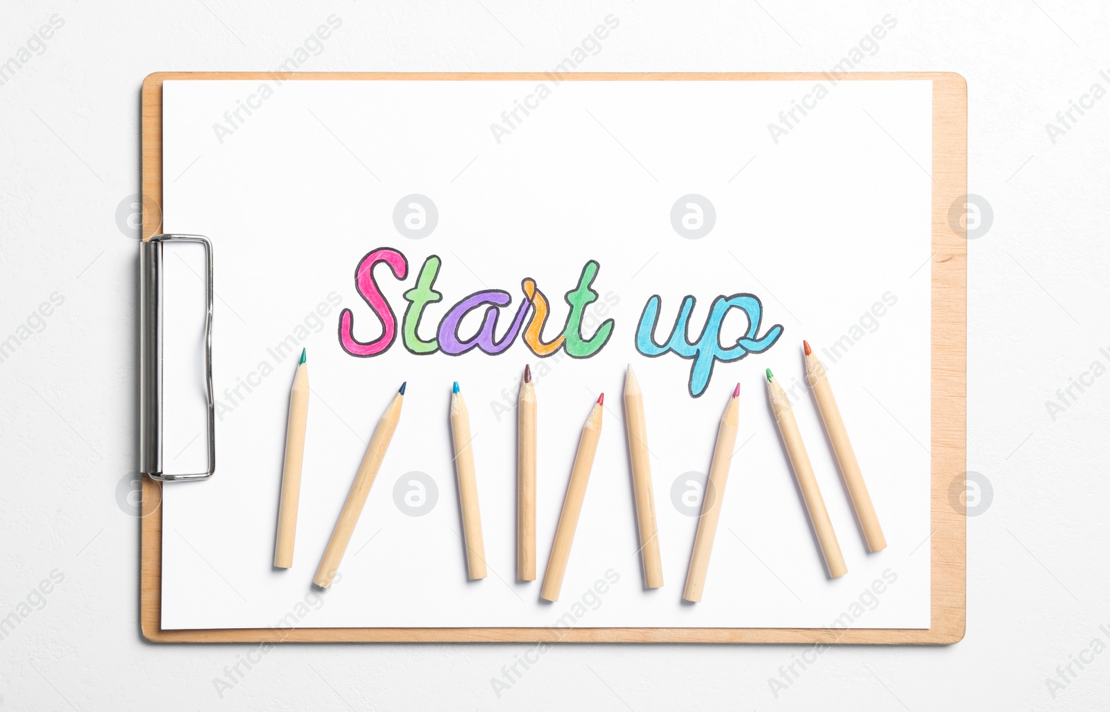 Photo of Clipboard with phrase Start Up and colorful pencils on white background, top view
