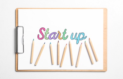 Photo of Clipboard with phrase Start Up and colorful pencils on white background, top view