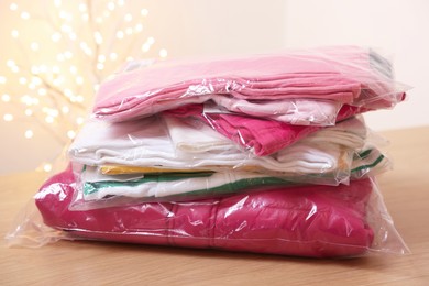 Photo of Stack of folded clothes in plastic bags on wooden table against beige background with blurred lights