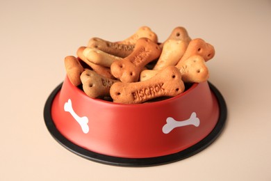 Photo of Bone shaped dog cookies in feeding bowl on beige table, closeup