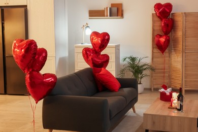 Photo of Romantic atmosphere. Burning candles, present, bottle of wine and glasses on wooden cabinet near sofa in room decorated for Valentine day