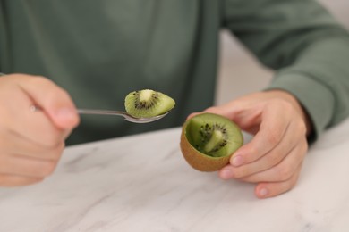 Photo of Boy eating tasty fresh kiwi with spoon at white marble table indoors, closeup