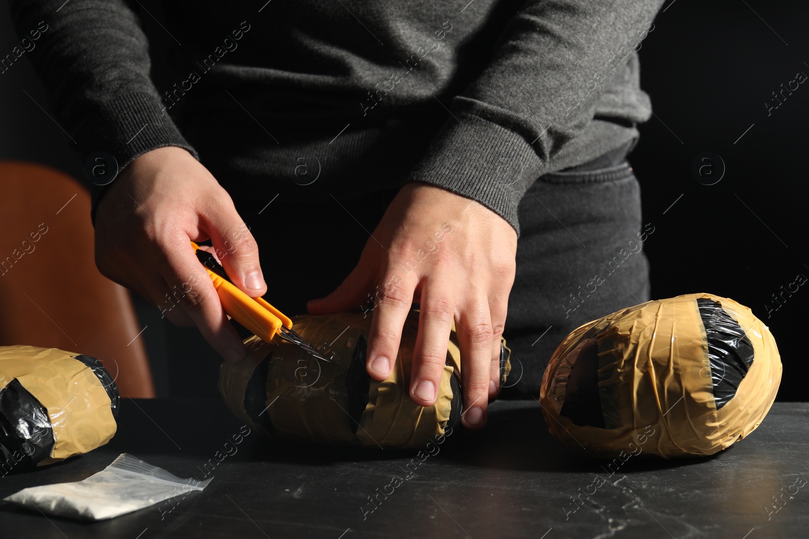 Photo of Smuggling and drug trafficking. Man opening package of narcotics with box cutter at dark table, closeup