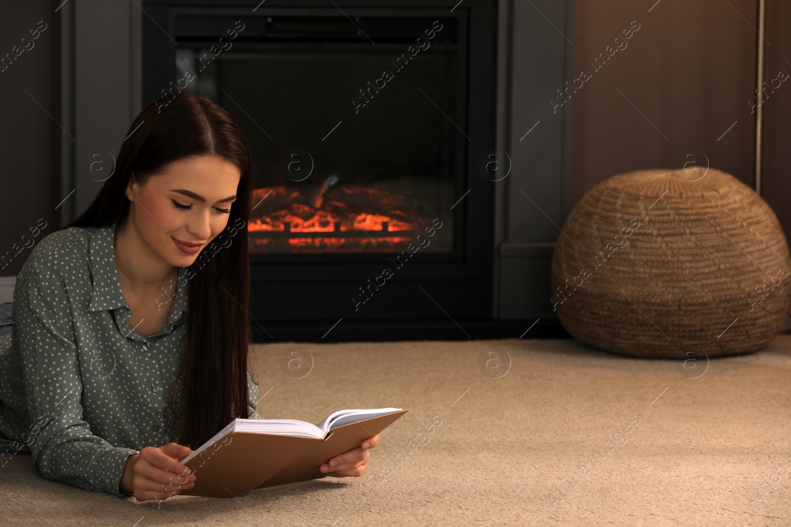 Photo of Beautiful young woman reading book on floor near fireplace at home. Space for text