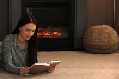 Photo of Beautiful young woman reading book on floor near fireplace at home. Space for text