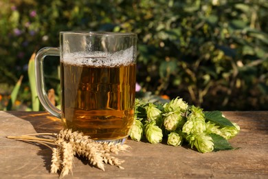 Photo of Mug with beer, fresh hops and ears of wheat on wooden table outdoors, space for text