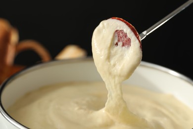 Photo of Dipping piece of sausage into fondue pot with melted cheese on black background, closeup