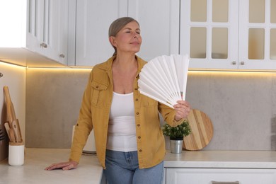 Photo of Menopause. Woman waving hand fan to cool herself during hot flash in kitchen