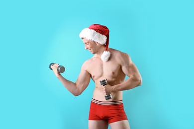 Photo of Young muscular man in Santa hat with dumbbells on color background