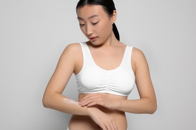 Photo of Beautiful young Asian woman applying body cream onto arm on light grey background