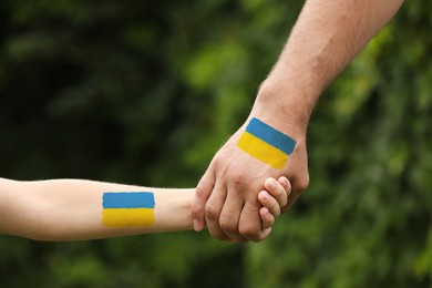 Image of Help for Ukraine. Man and child holding hands with drawings of Ukrainian flag outdoors, closeup