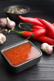 Photo of Spicy chili sauce, garlic, peppers and peppercorns on black wooden table, closeup