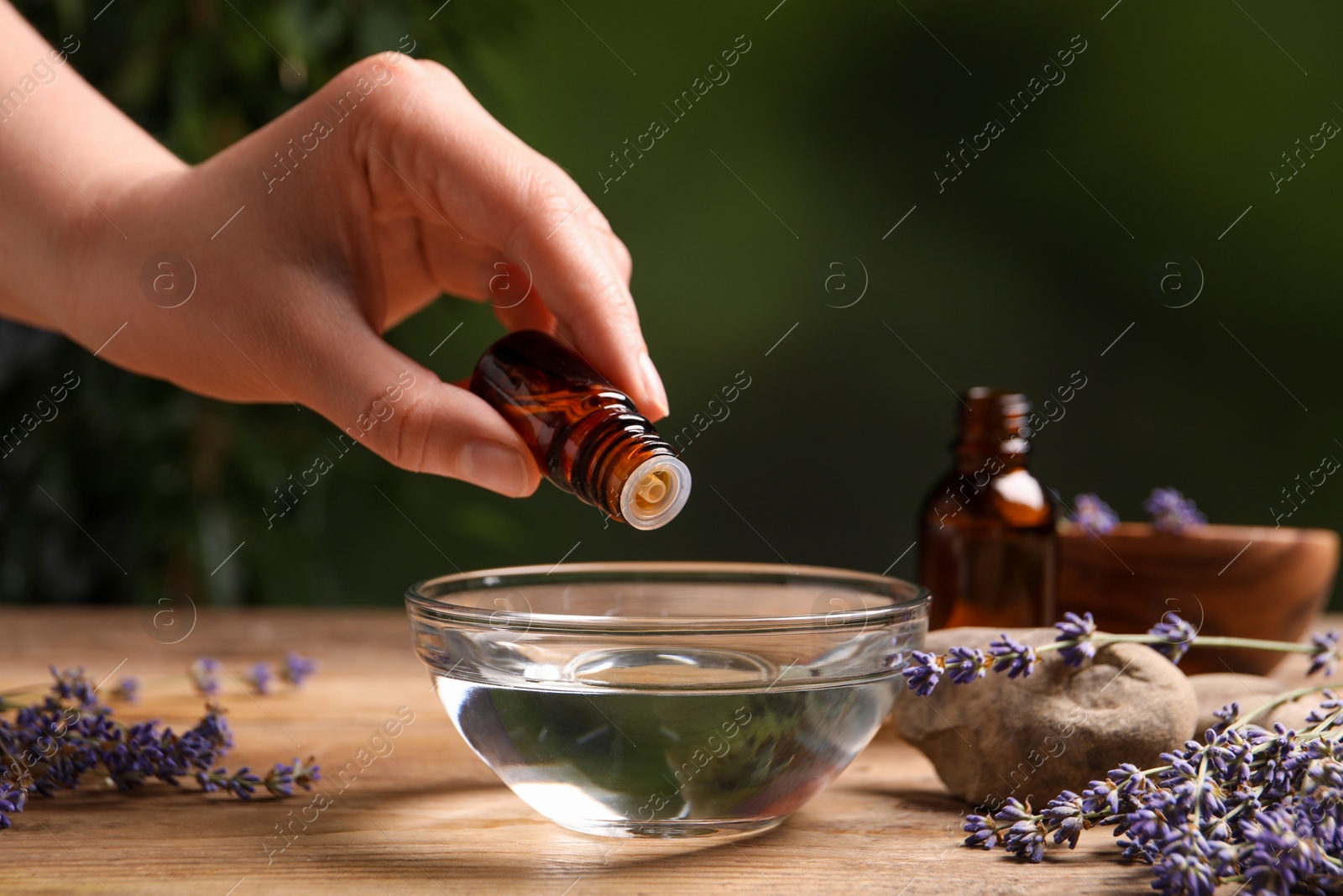 Photo of Woman dripping lavender essential oil from bottle into bowl at wooden table, closeup