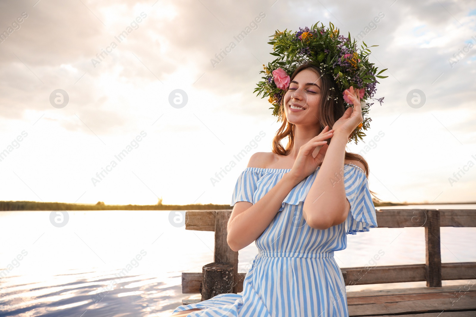 Photo of Young woman wearing wreath made of beautiful flowers on pier near river