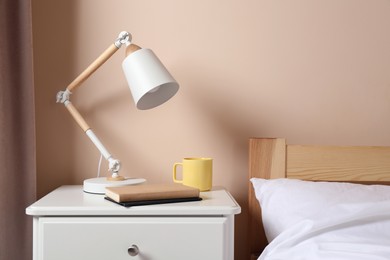 Stylish modern desk lamp, books and cup of drink on white nightstand in bedroom