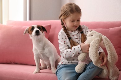 Photo of Cute little girl with her dog and toy bunny on sofa indoors. Childhood pet