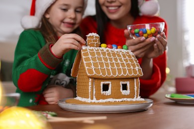 Photo of Mother and daughter decorating gingerbread house at table indoors, closeup