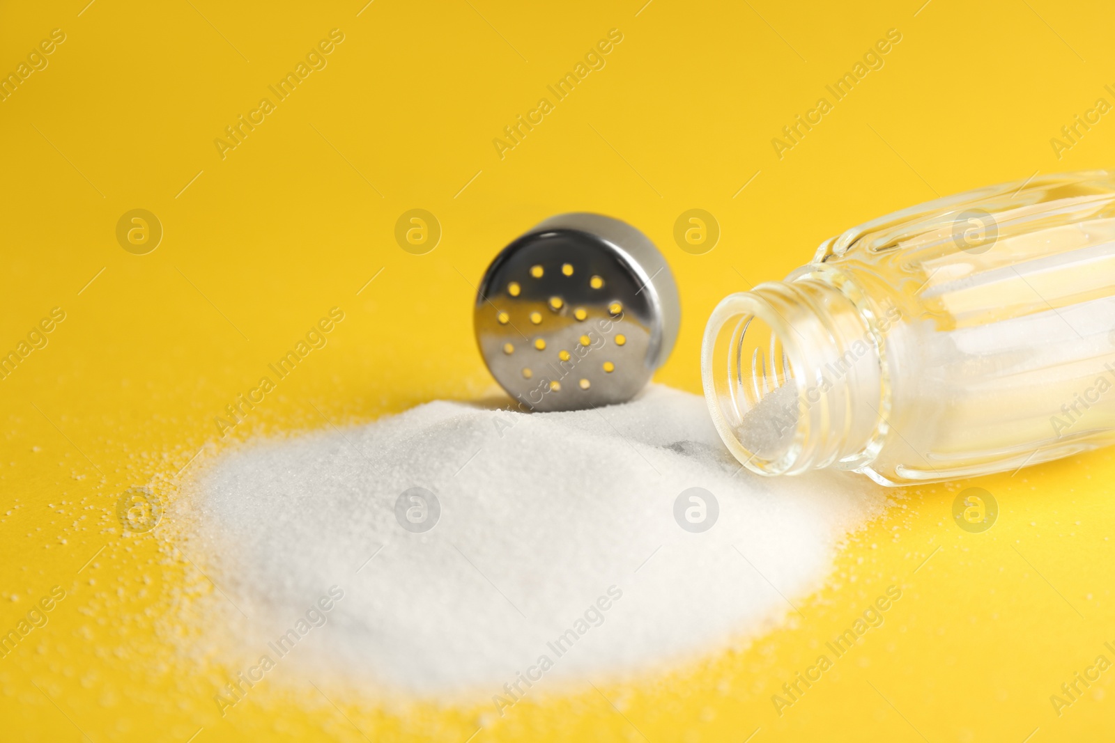 Photo of Scattered salt and shaker on yellow background, closeup