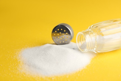 Photo of Scattered salt and shaker on yellow background, closeup