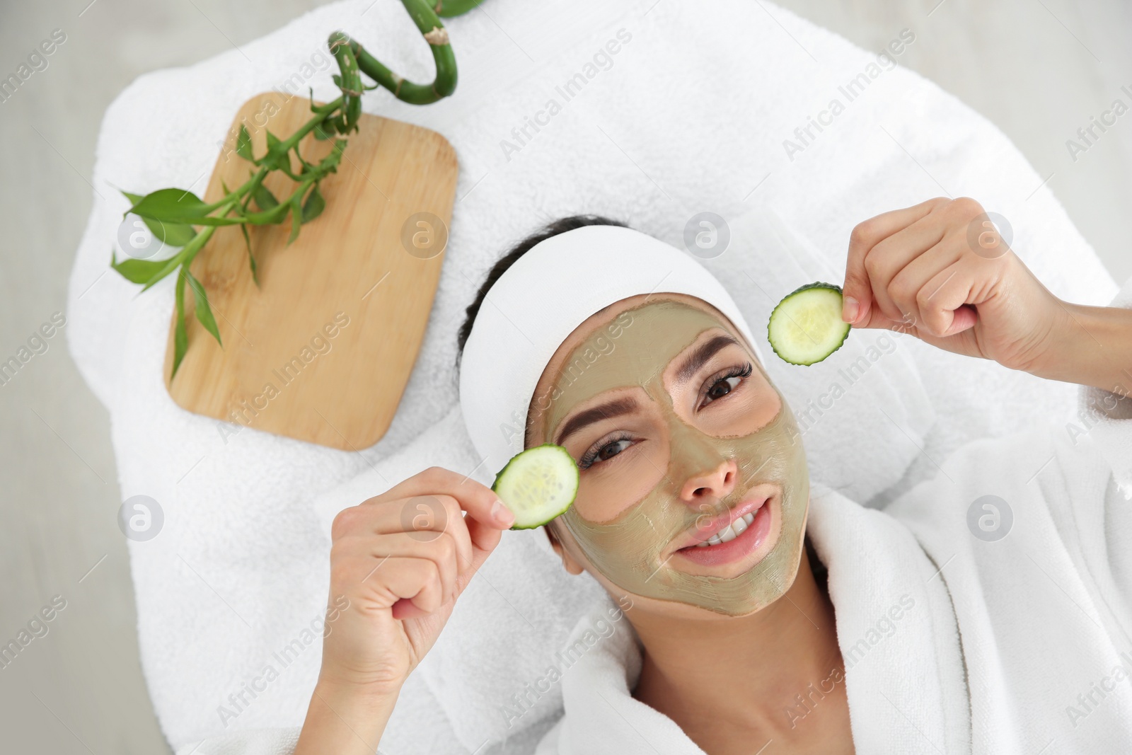 Photo of Pretty woman with clay mask on her face holding cucumber slices in spa salon, above view